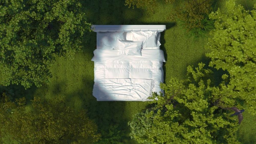 A bed with white linens stands on the grass in the forest among the trees