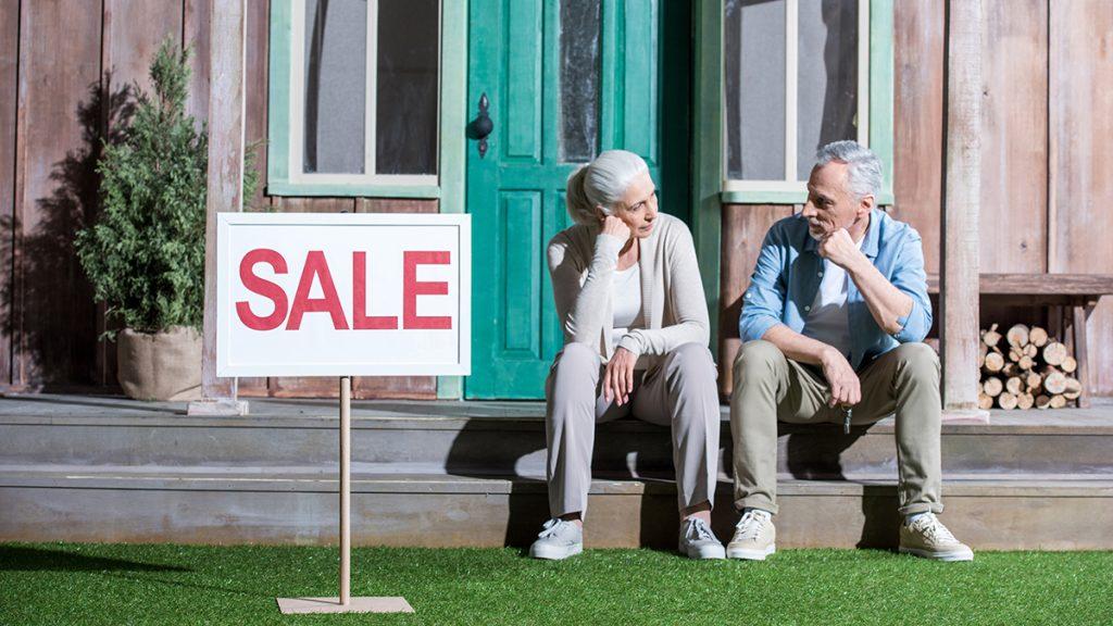 Upset senior couple sitting on porch next to sale sign